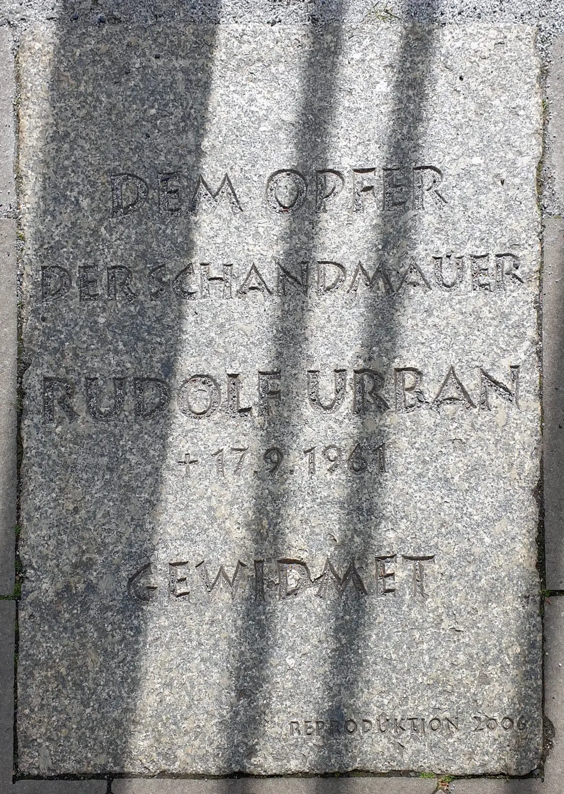 Granite slab in the pavement engraved "Dem Opfer der Schandmauer Rudolf Urban † 17.9.1961 gewidmet", crossed by shadows