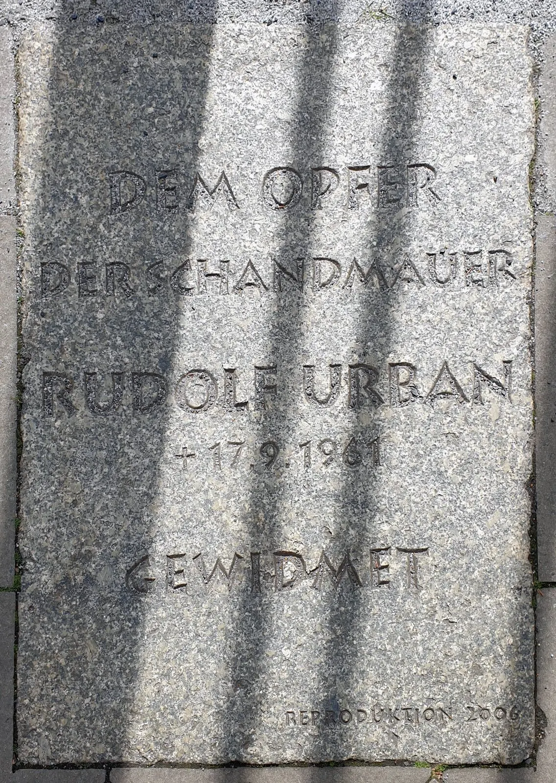 Granite slab in the pavement engraved "Dem Opfer der Schandmauer Rudolf Urban † 17.9.1961 gewidmet", crossed by shadows