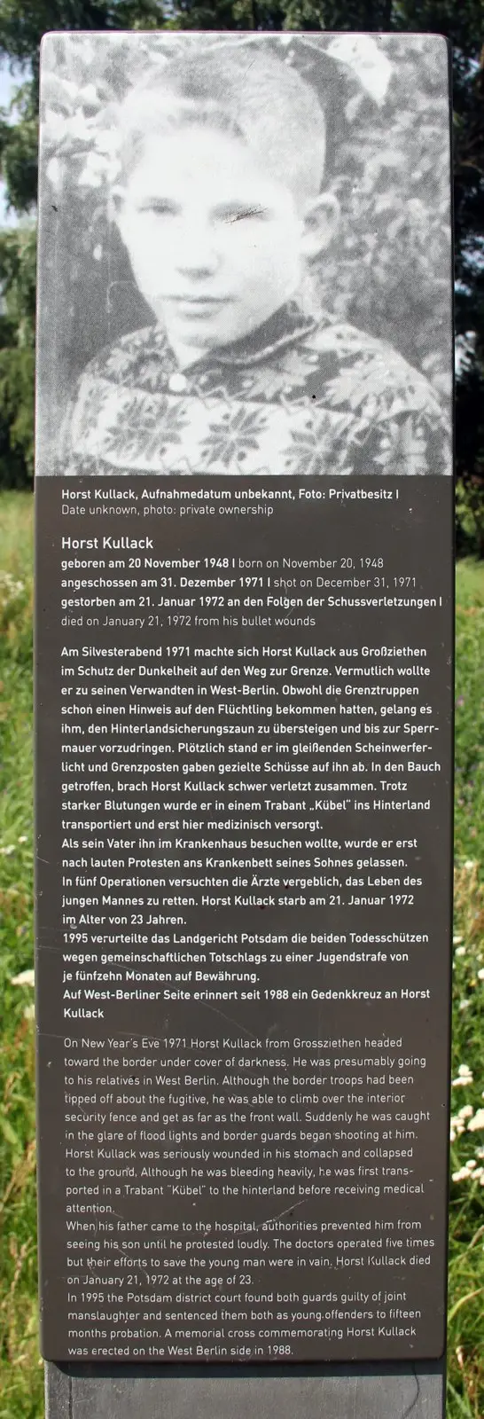 Dark stele topped by a black-and-white photograph of a boy in a patterned sweater, above printed German and English text