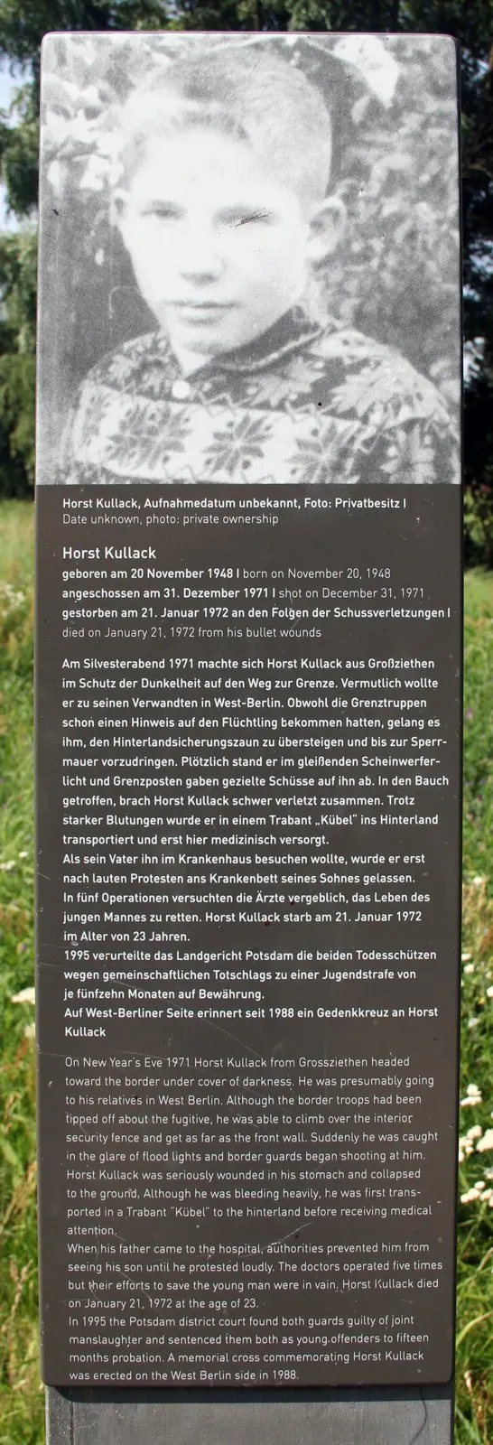 Dark stele topped by a black-and-white photograph of a boy in a patterned sweater, above printed German and English text