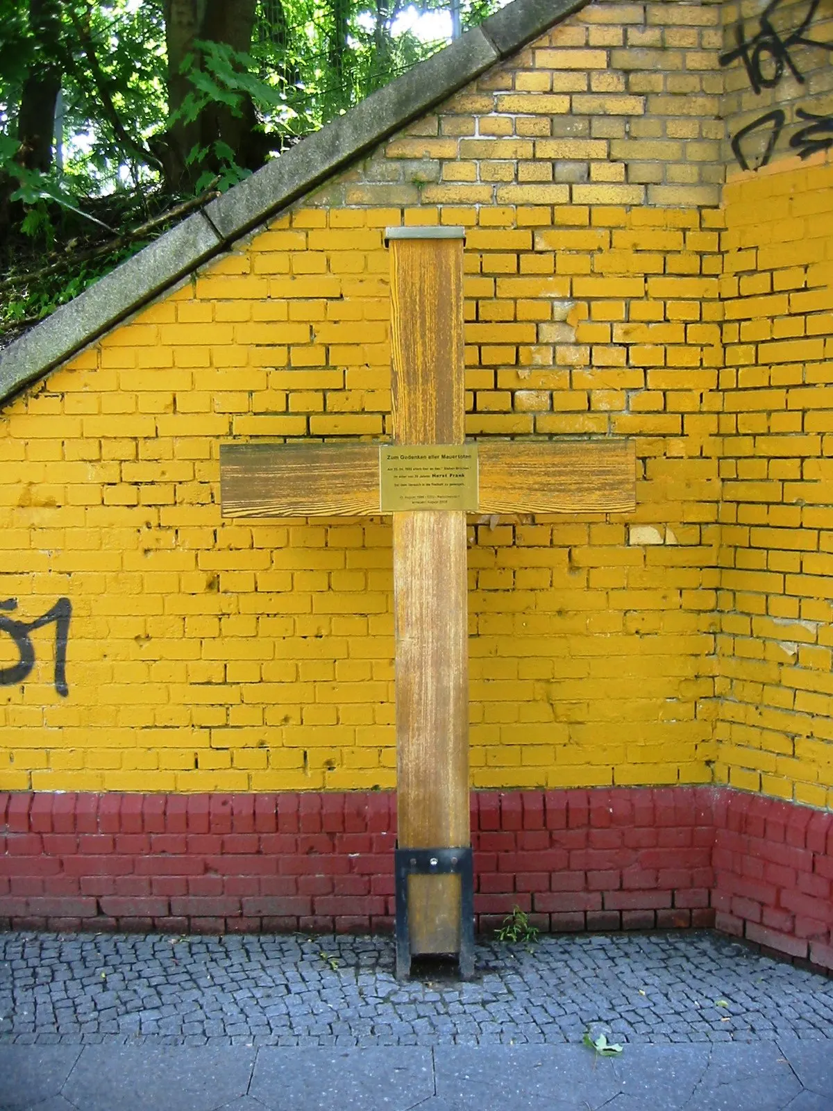 Wooden cross with a small plaque against a yellow and red painted brick wall