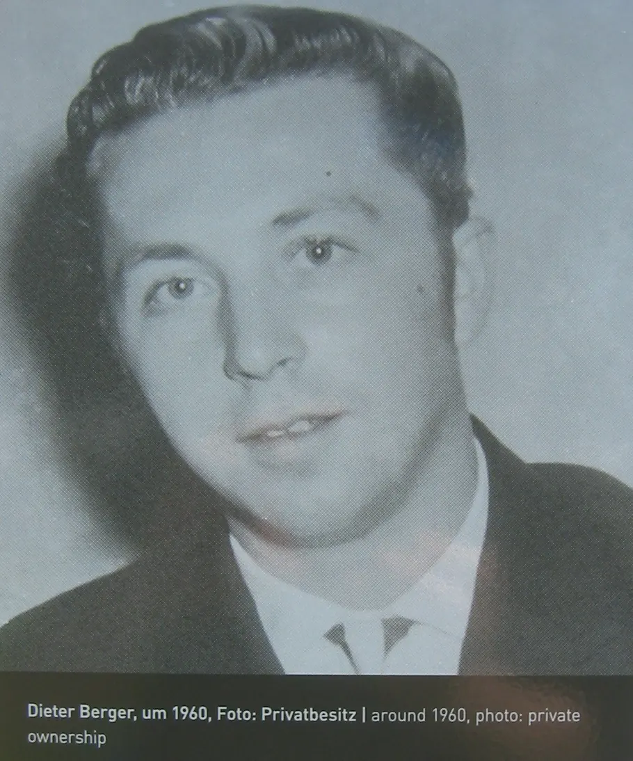 Black-and-white portrait of a young man with wavy dark hair in a jacket, white shirt and tie, with a printed caption below