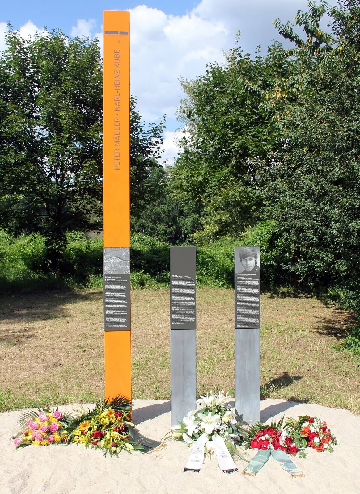 Tall orange column inscribed 'Peter Mädler · Karl-Heinz Kube' beside two grey posts with text panels, flowers and wreaths on sand at the base