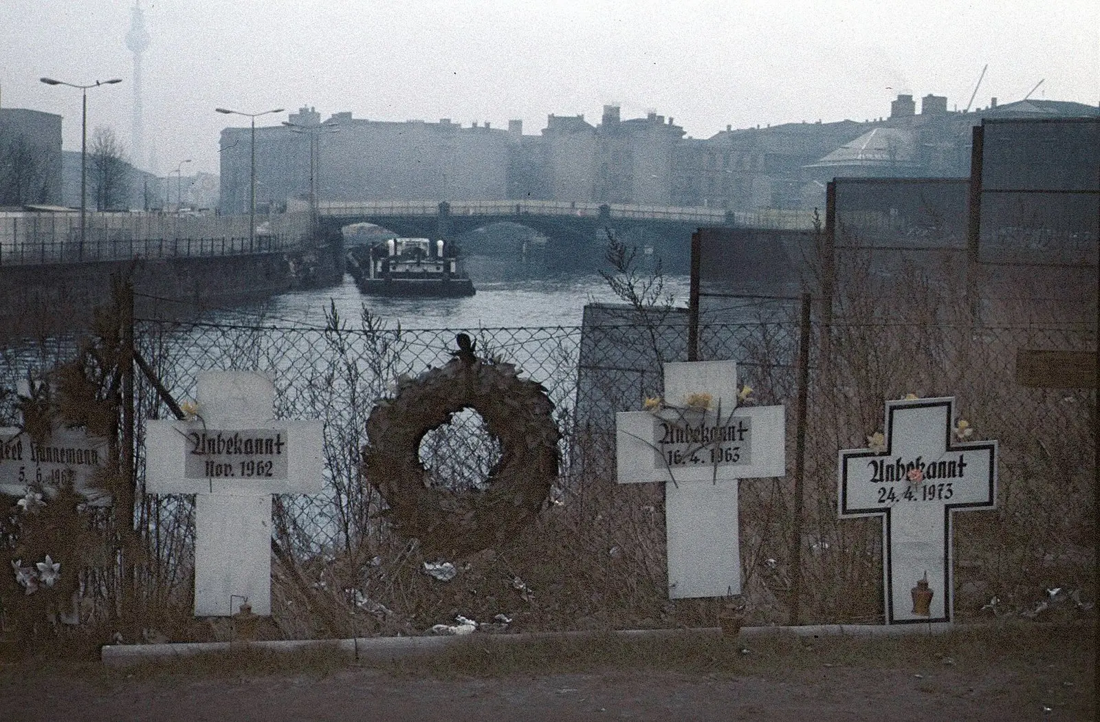 White crosses and a wreath on a wire fence above the river Spree, with a bridge and a barge in the background; the partly visible cross at the left edge bears the name Hannemann and the date 5.6.1962