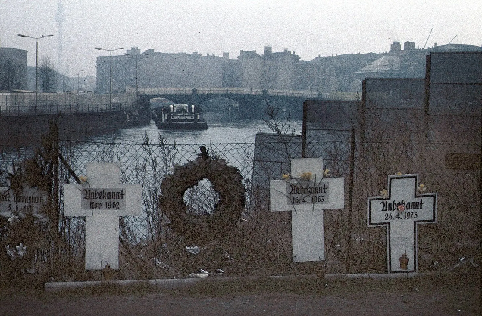 White crosses and a wreath on a wire fence above the river Spree, with a bridge and a barge in the background; the partly visible cross at the left edge bears the name Hannemann and the date 5.6.1962