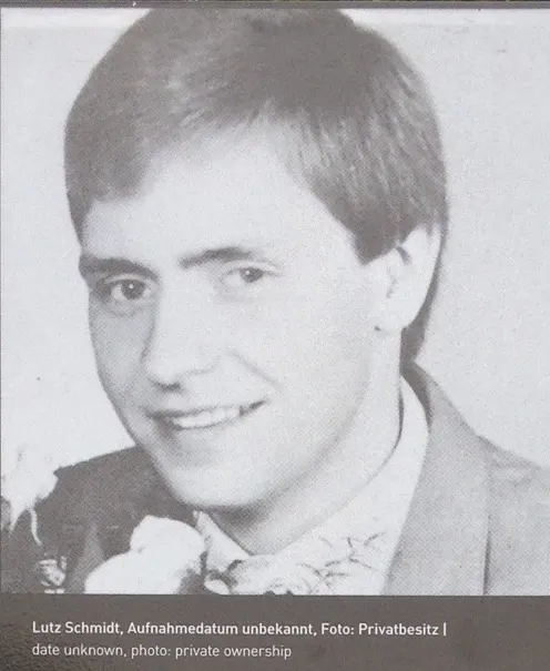 Black-and-white portrait of a smiling young man with short hair, in a jacket with a flower at the lapel, with a caption strip below
