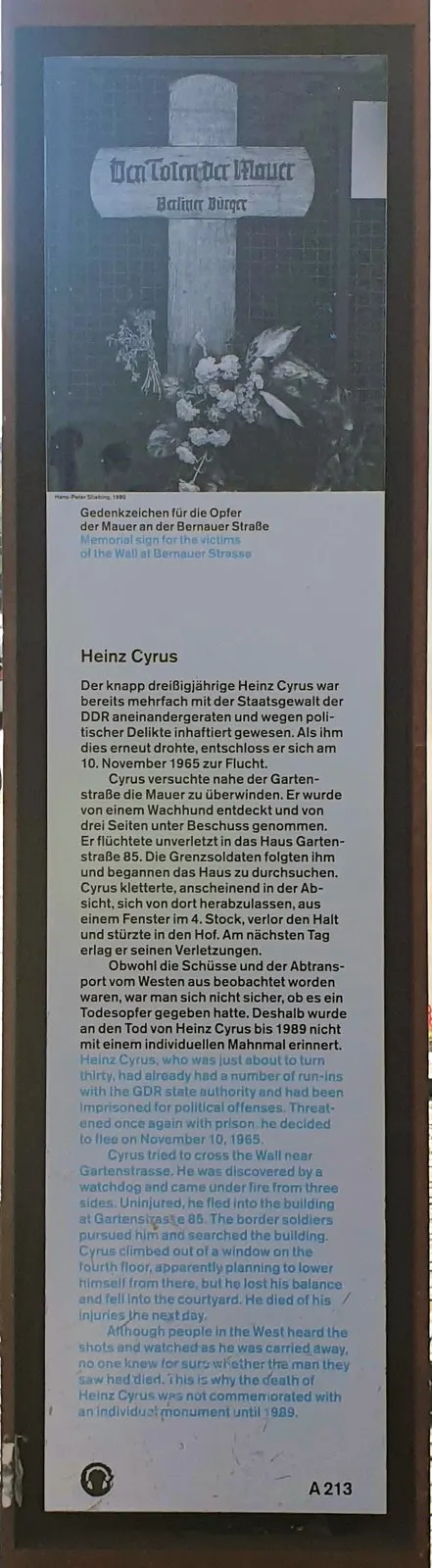 Tall panel with a black-and-white photograph of a wooden cross with flowers, above German and English text headed "Heinz Cyrus"