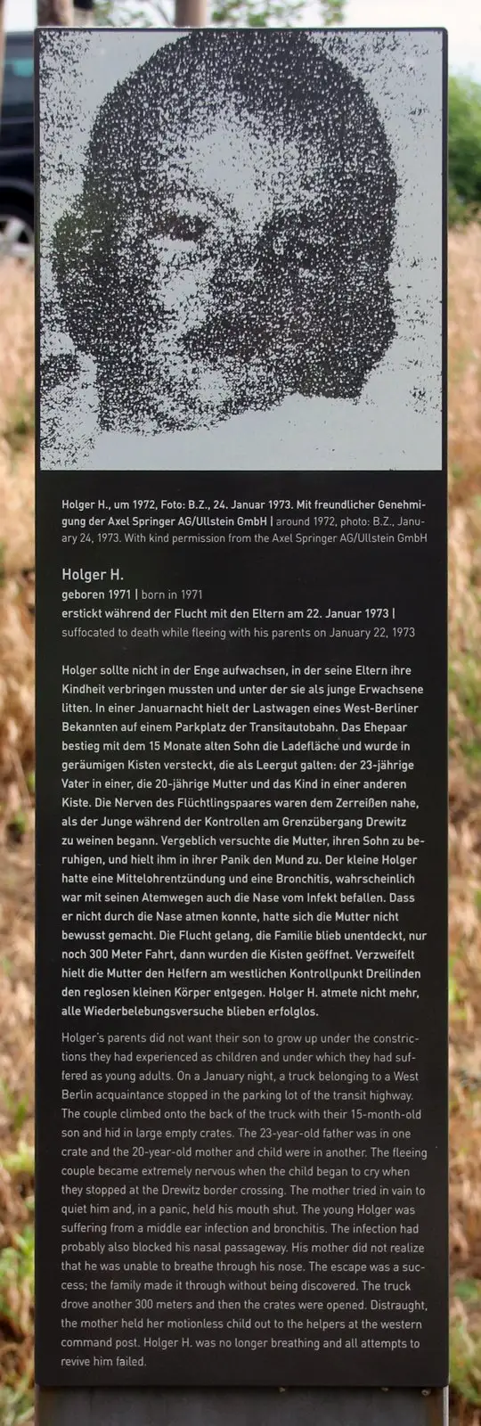 Dark stele topped by a coarse-grained black-and-white photograph of a small child's face, above printed German and English text, in dry grass