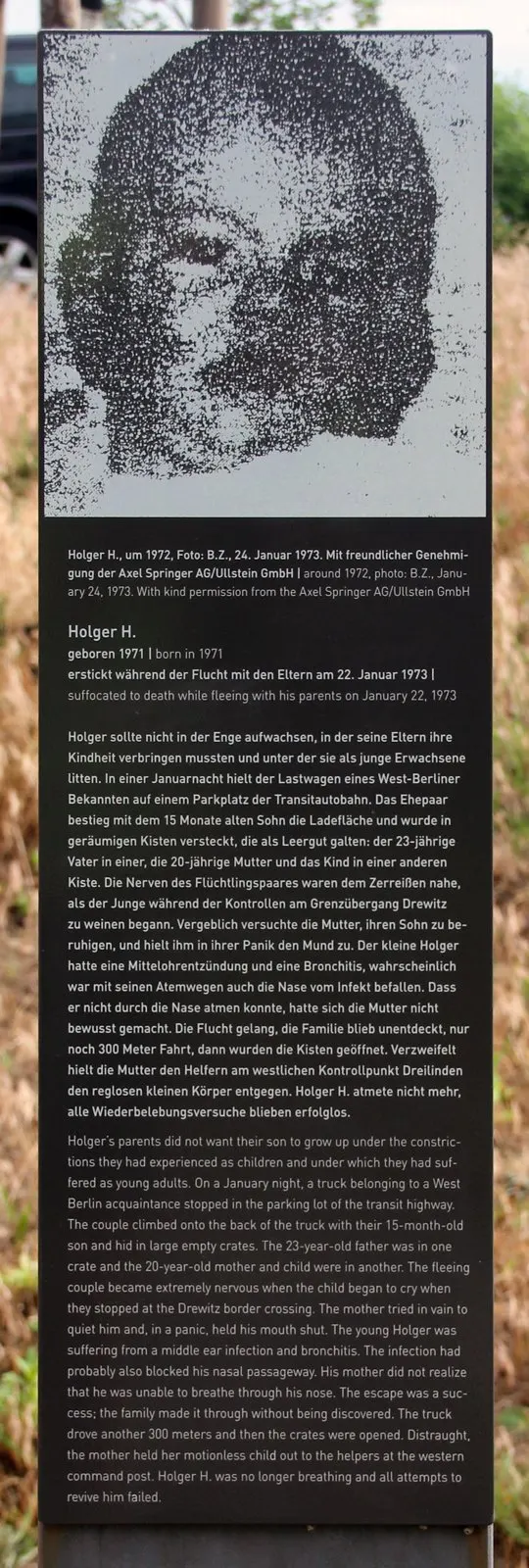 Dark stele topped by a coarse-grained black-and-white photograph of a small child's face, above printed German and English text, in dry grass