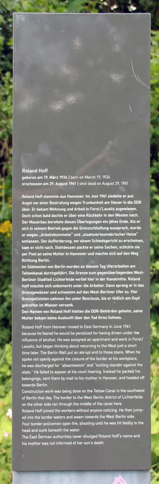 Dark grey information stele with German and English text about Roland Hoff, standing among green shrubs