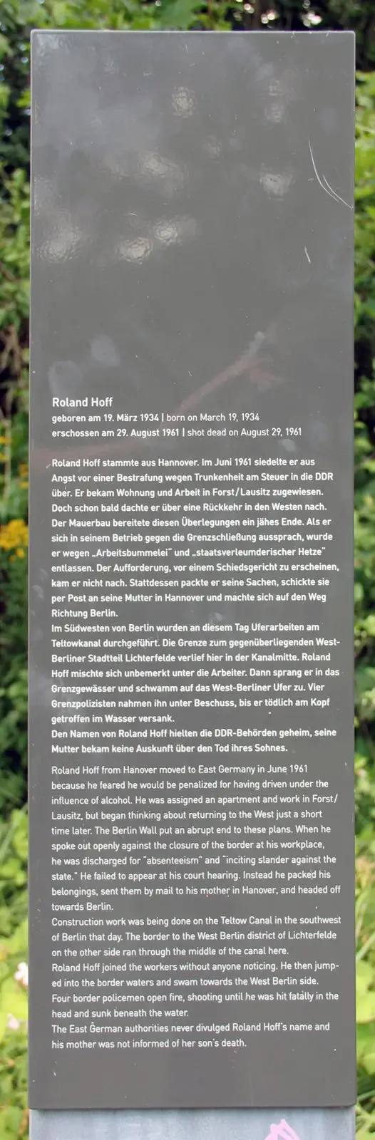 Dark grey information stele with German and English text about Roland Hoff, standing among green shrubs