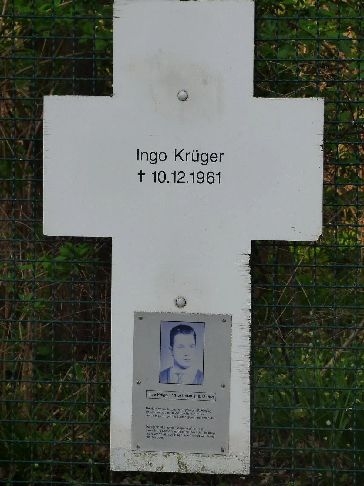 Close-up of a white cross on a green wire fence bearing the name Ingo Krüger, with a small metal plate showing a portrait photo and text below