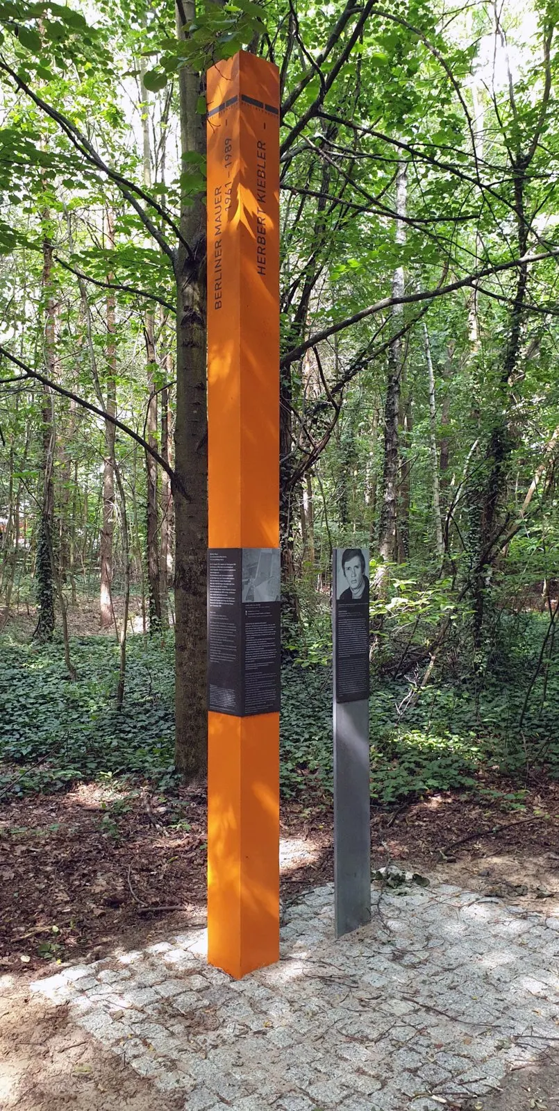 Tall orange column bearing his name standing in a wood, next to a slim grey post with a portrait photograph and text panel