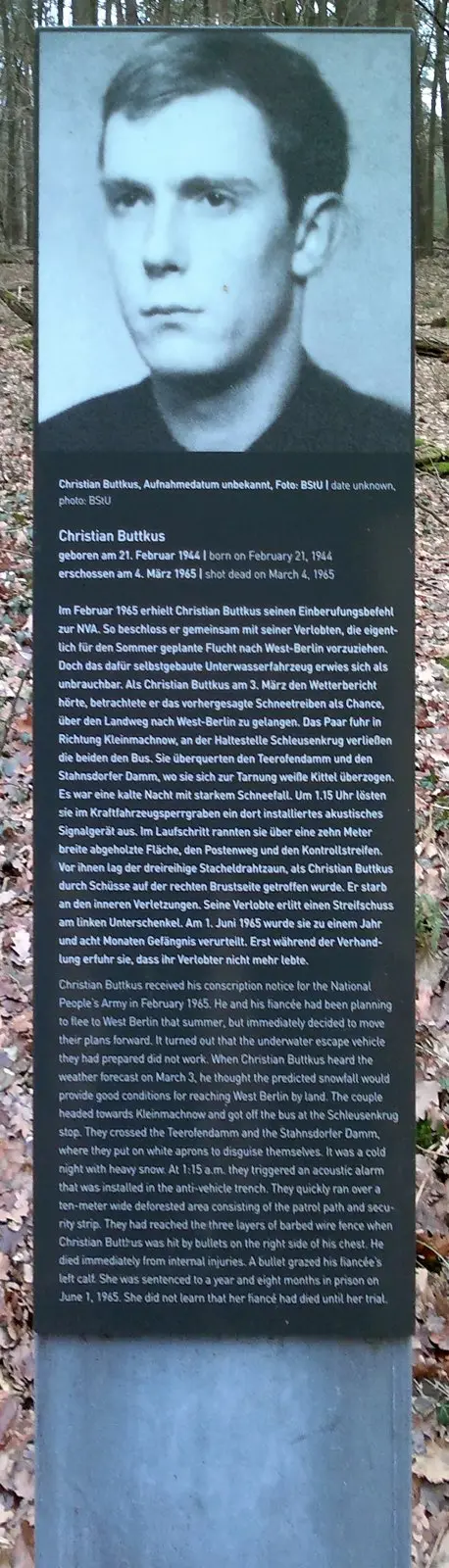 Dark stele with a black-and-white portrait of a young man above a German and English text headed 'Christian Buttkus', in bare woodland