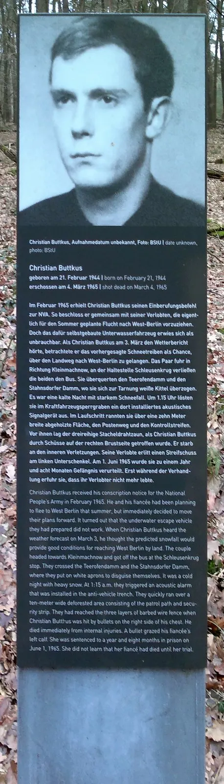 Dark stele with a black-and-white portrait of a young man above a German and English text headed 'Christian Buttkus', in bare woodland
