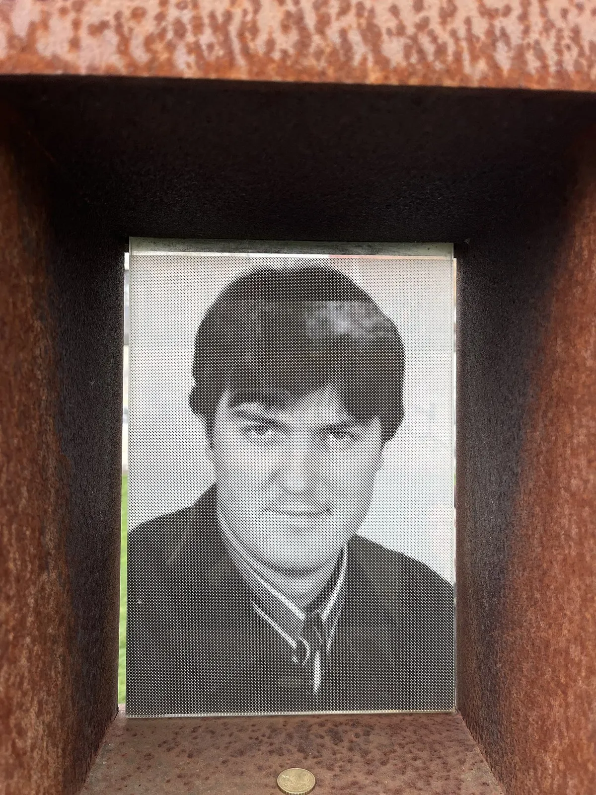 Black-and-white photograph of a young man with dark hair, jacket and tie, set in a rusted steel niche