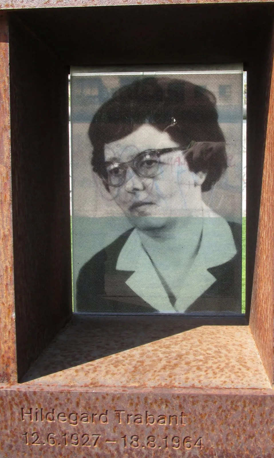 Black-and-white portrait of a woman with glasses and short dark hair, set in a rusted steel niche engraved 'Hildegard Trabant 12.6.1927 – 18.8.1964'