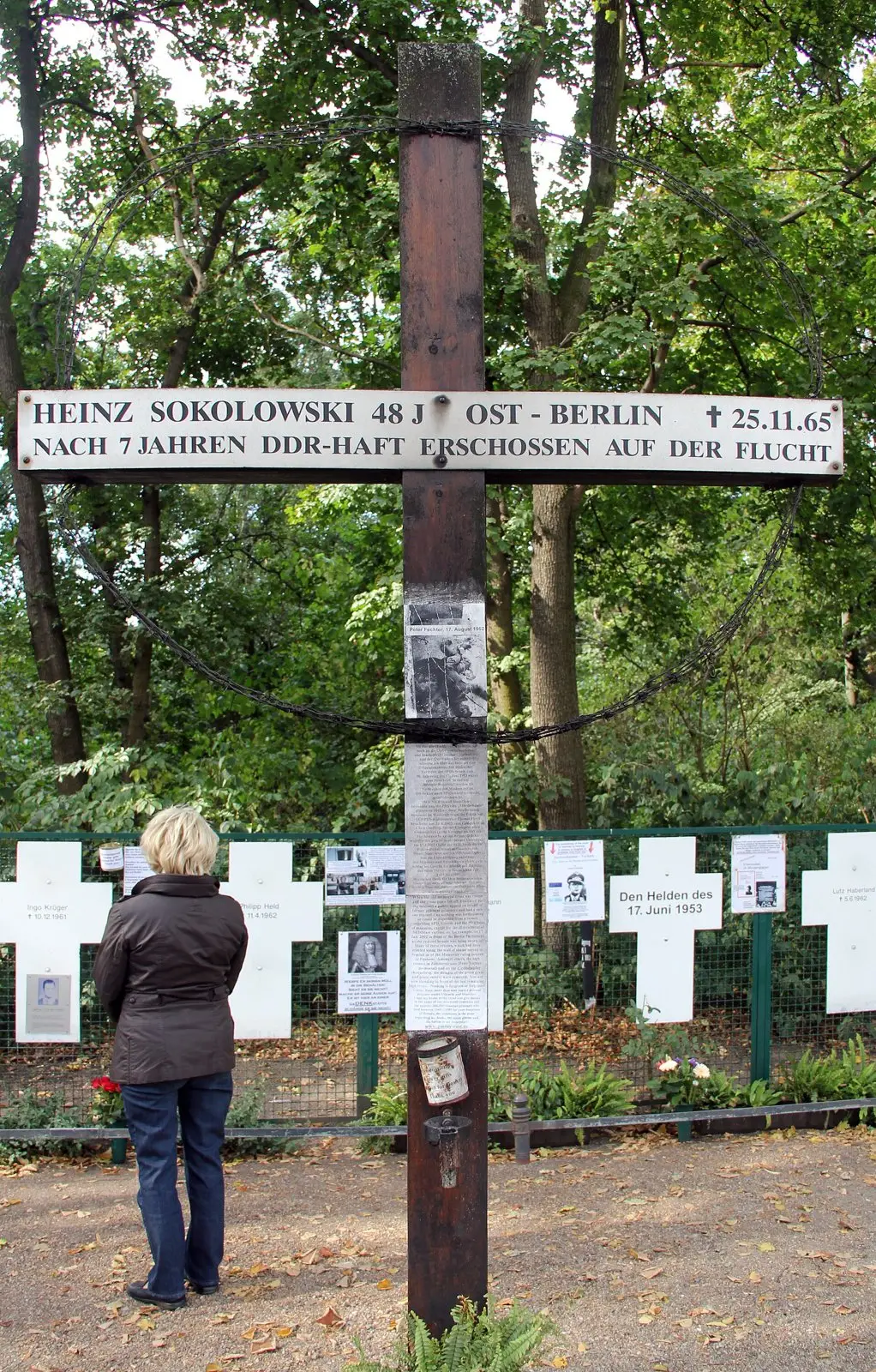 Tall dark wooden cross with a white sign reading "Heinz Sokolowski 48 J. Ost-Berlin † 25.11.65 – nach 7 Jahren DDR-Haft erschossen auf der Flucht", in front of white crosses and trees; a woman stands looking at it