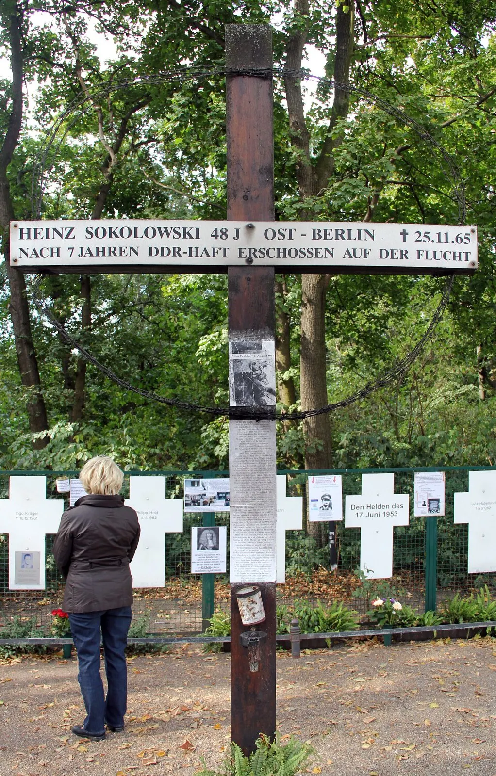 Tall dark wooden cross with a white sign reading "Heinz Sokolowski 48 J. Ost-Berlin † 25.11.65 – nach 7 Jahren DDR-Haft erschossen auf der Flucht", in front of white crosses and trees; a woman stands looking at it