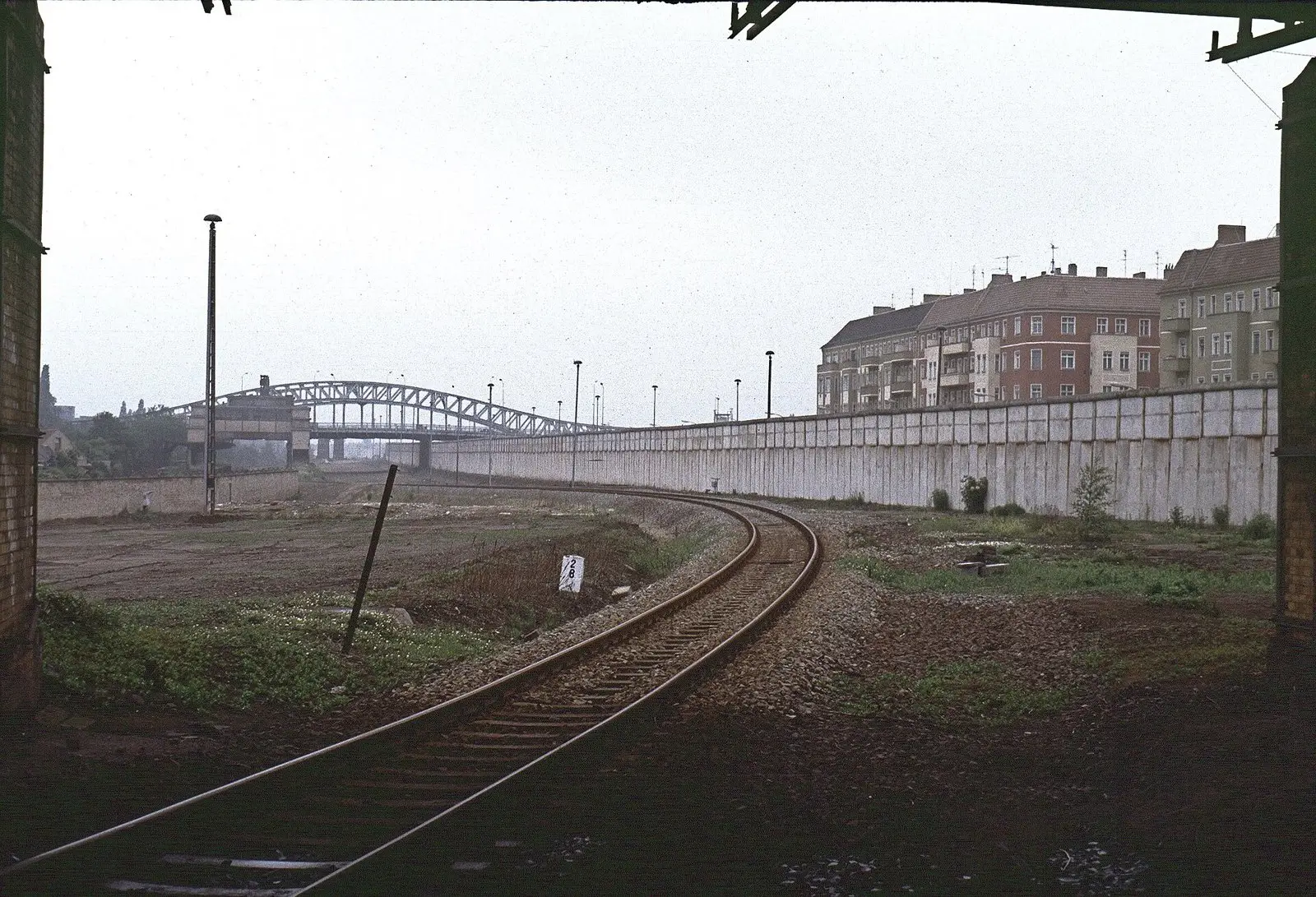 Colour photograph of a single railway track curving between a grassy strip and a long white concrete wall towards a steel arch bridge; apartment buildings stand behind the wall on the right.