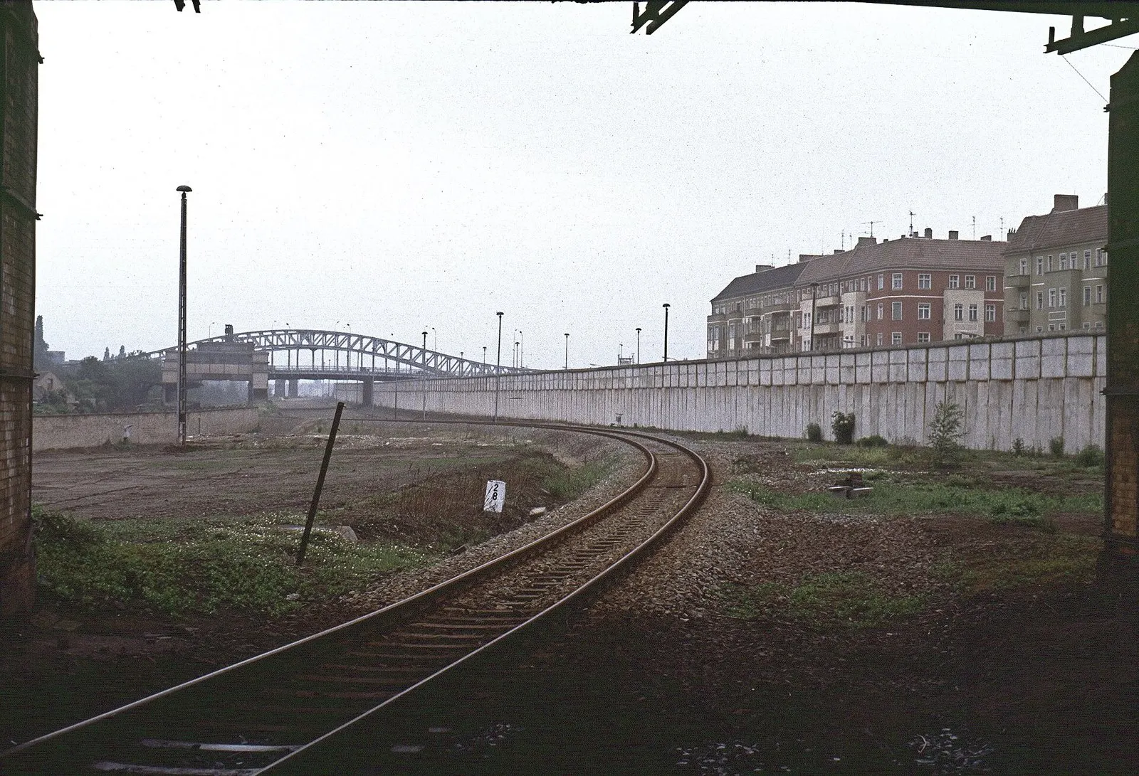 Colour photograph of a single railway track curving between a grassy strip and a long white concrete wall towards a steel arch bridge; apartment buildings stand behind the wall on the right.