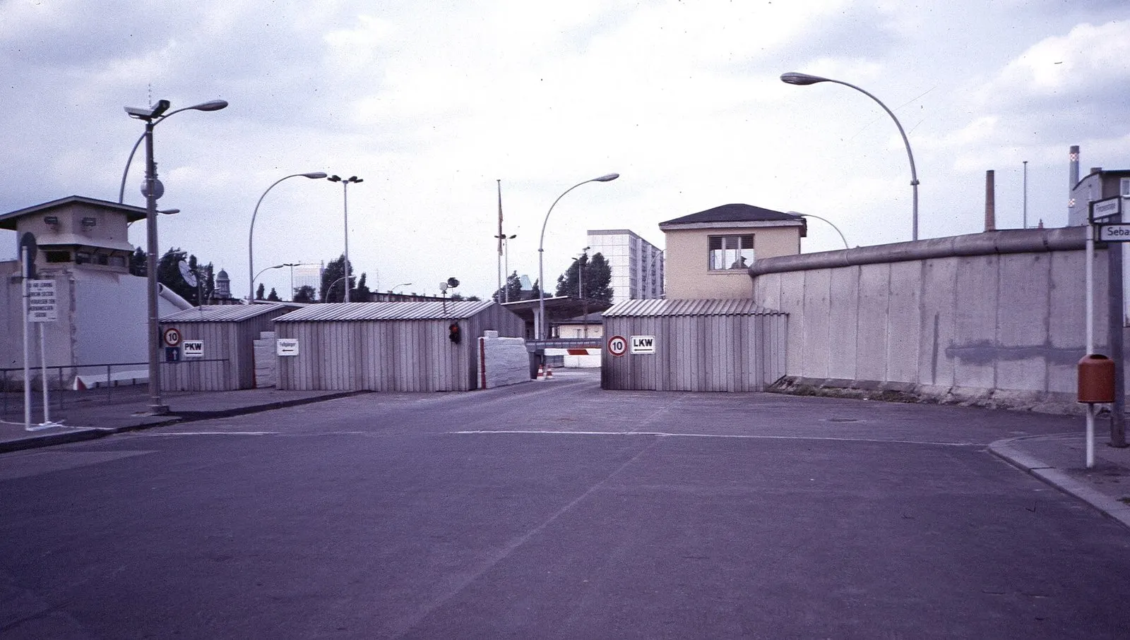 Colour photograph of a border checkpoint: an empty paved roadway leads to low corrugated huts with signs for cars (PKW) and lorries (LKW); a long concrete wall runs along the right, curved street lamps and a small watch building stand on the left.