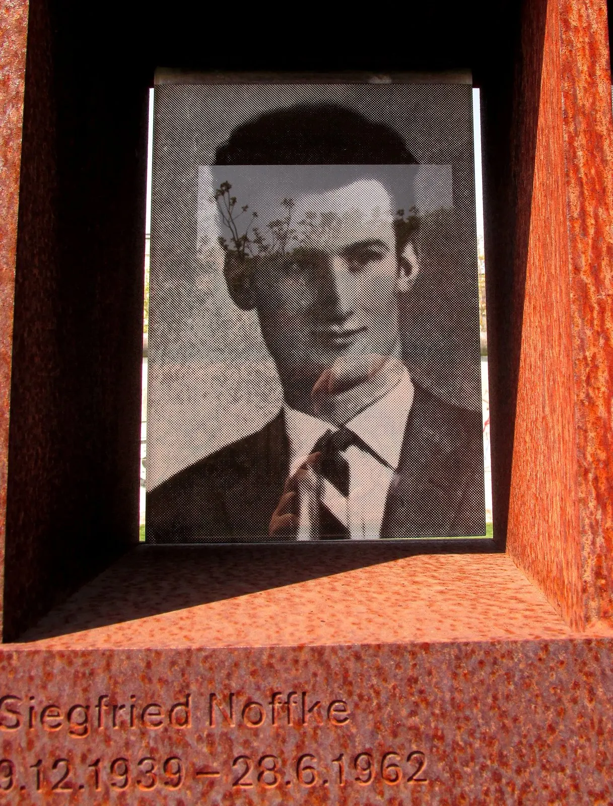 Black-and-white photograph of a young man in a suit and tie behind glass in a rusted steel niche, his name engraved below