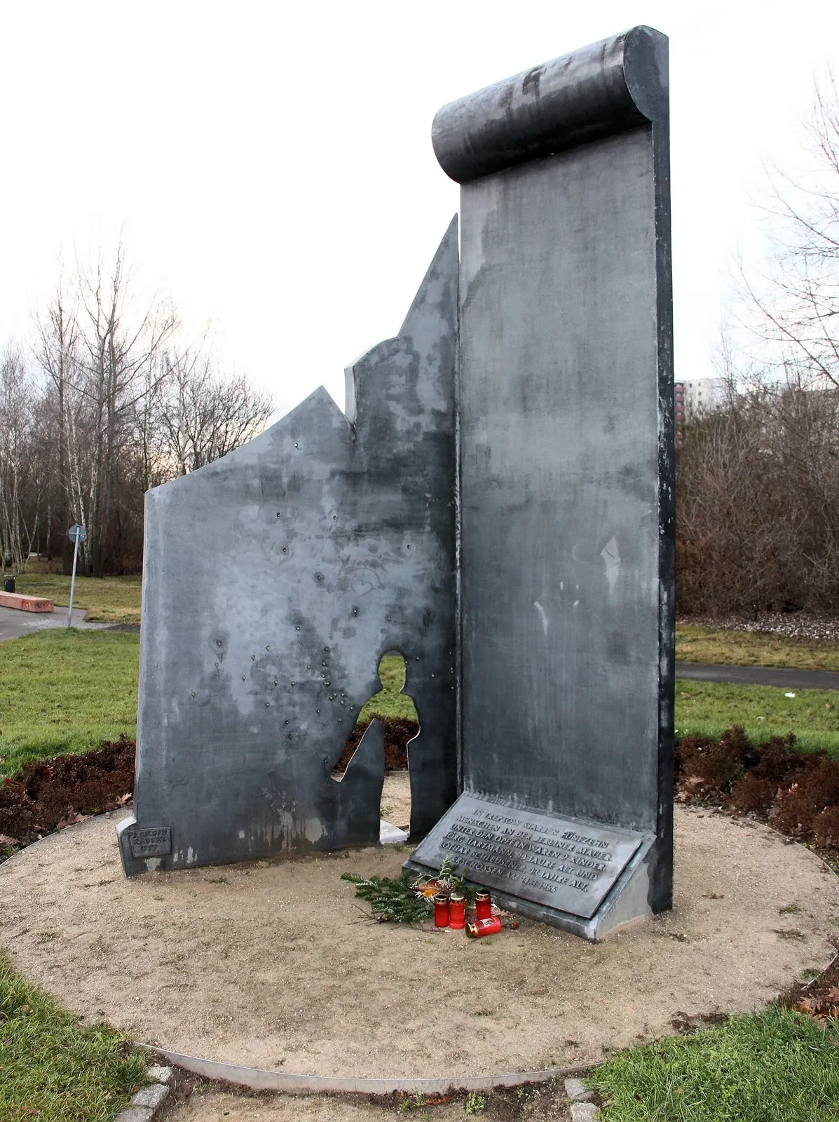Dark grey metal sculpture of a broken wall with bullet holes and a child-shaped opening, beside an upright slab with an inscribed plaque, with flowers and candles at its foot