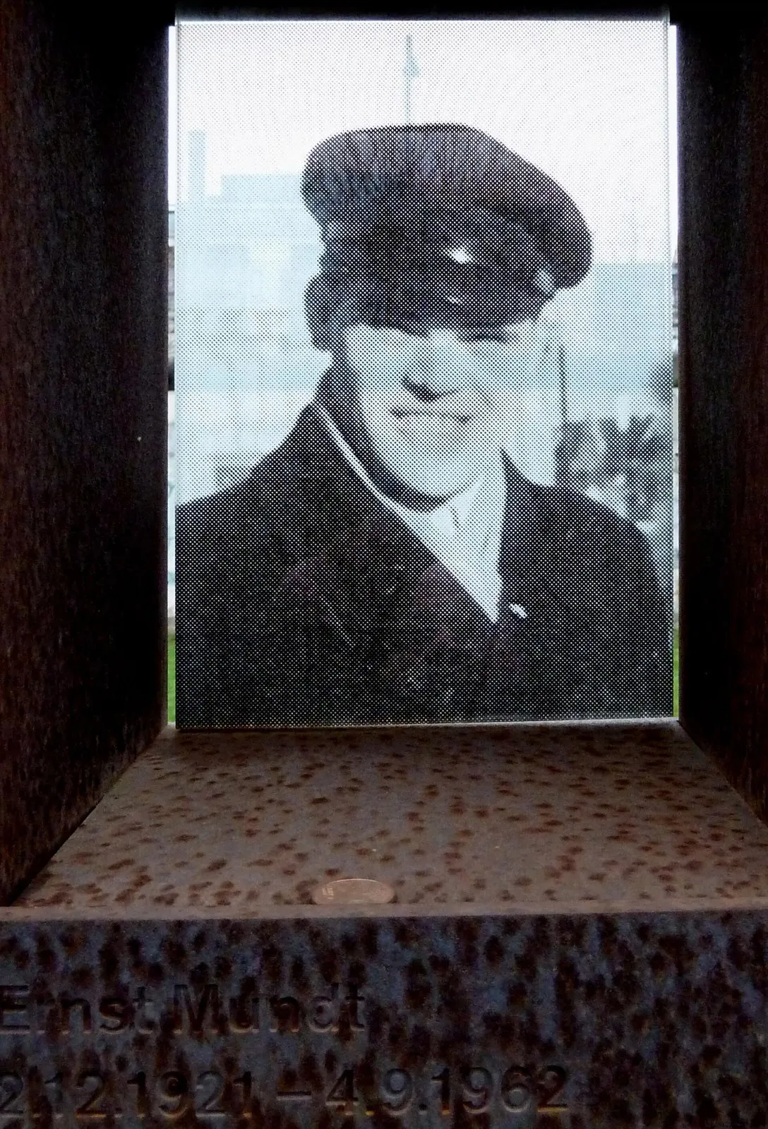 Black-and-white photograph of a man in a peaked cap and dark coat behind glass in a steel niche, his name engraved below