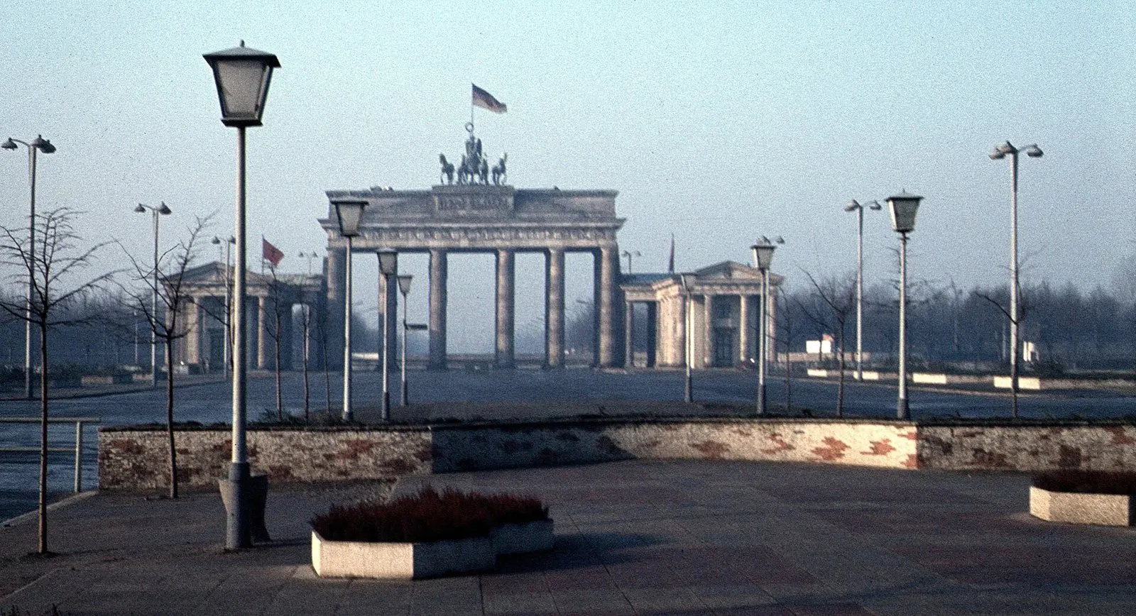 Colour photograph of the Brandenburg Gate with its quadriga at the far end of a wide, empty square lined with lamp posts and young trees; a low brick wall and planters in the foreground under a hazy blue sky.