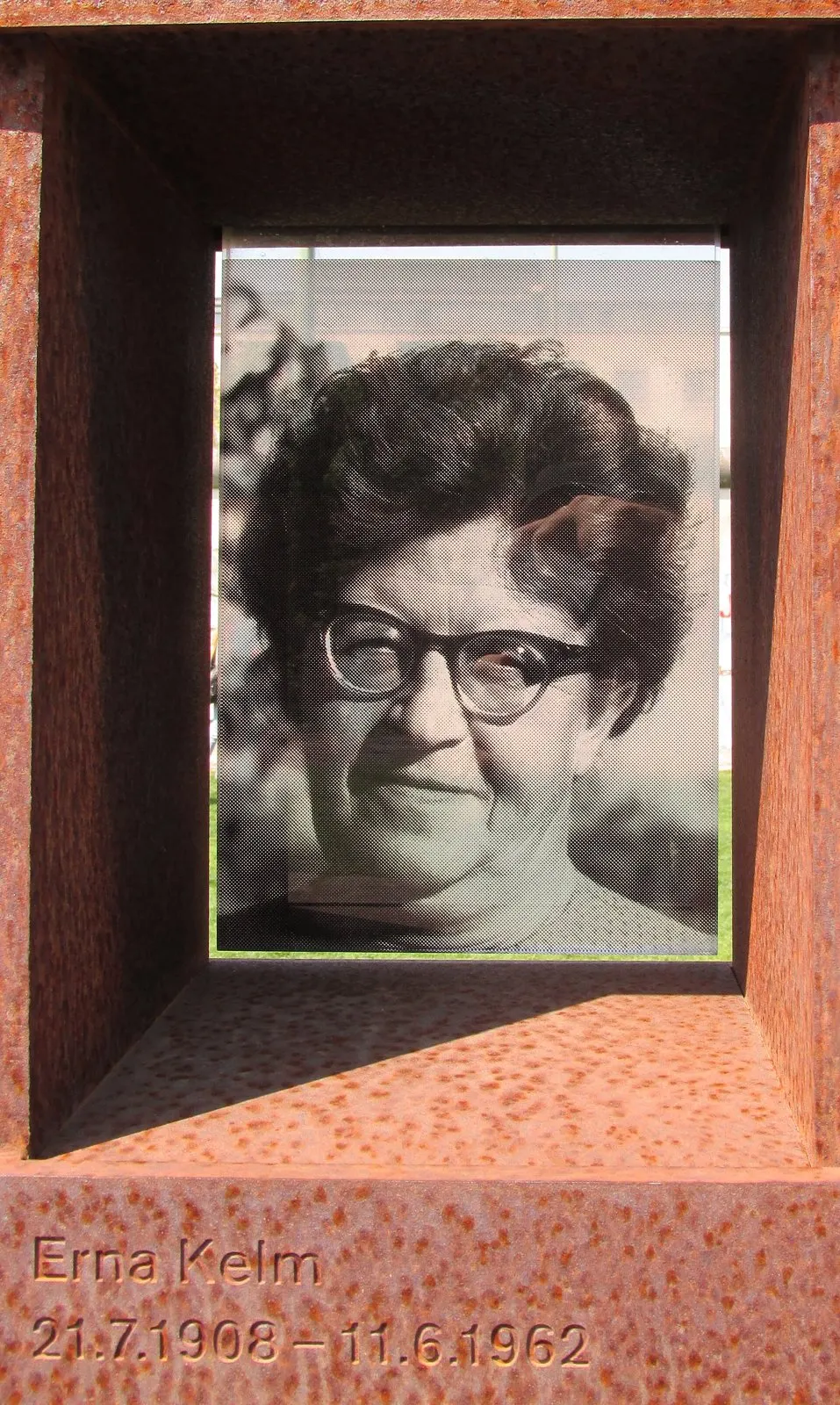 Black-and-white photograph of an older woman with glasses and curly hair behind glass in a rusted steel niche, her name engraved below