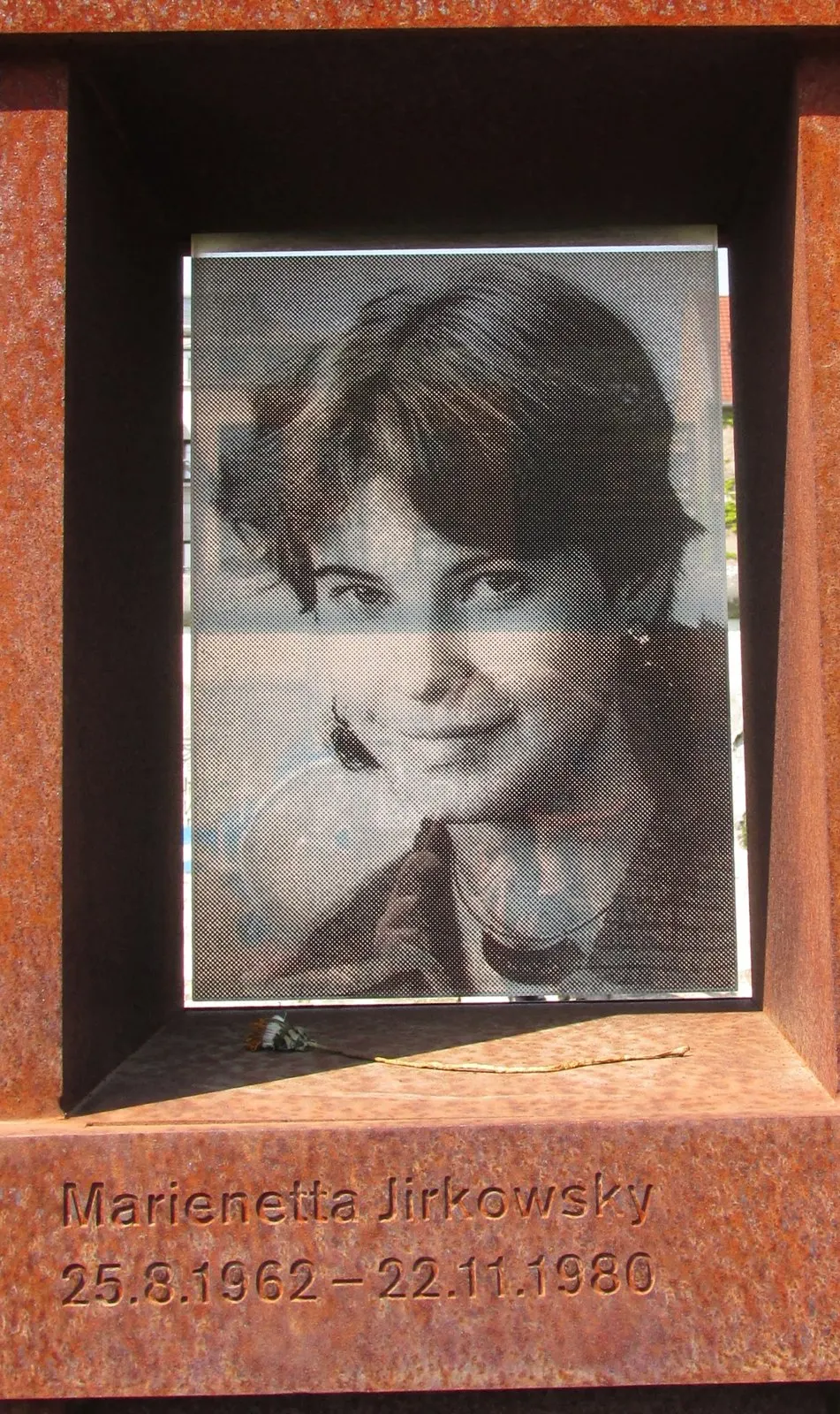 Black-and-white photograph of a smiling young woman with short dark hair, set in a rusted steel niche engraved with her name and dates