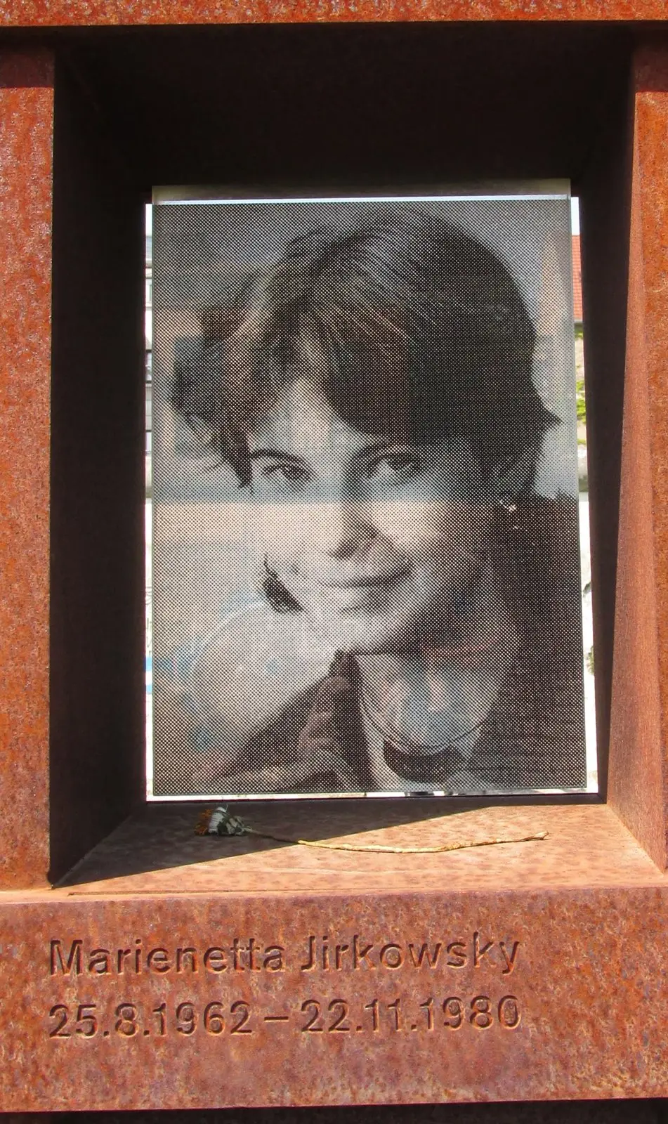 Black-and-white photograph of a smiling young woman with short dark hair, set in a rusted steel niche engraved with her name and dates