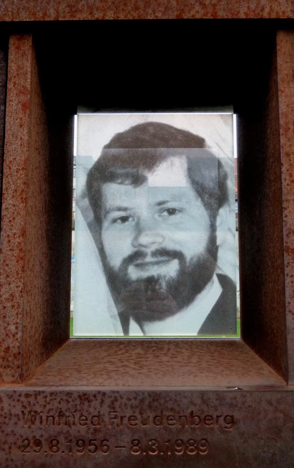 Black-and-white photograph of a bearded man in a jacket and tie, set in a rusted steel niche engraved with his name and dates