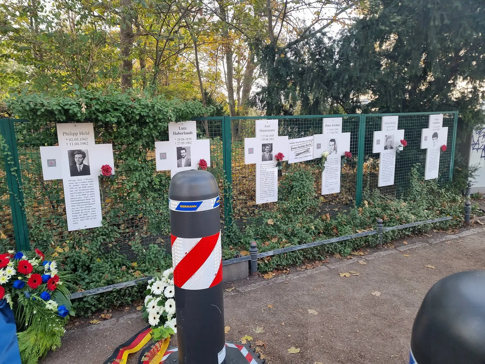 Row of six white crosses on a green fence under trees, each with a name, dates, a black-and-white portrait and a short text; the second cross from the right reads Dieter Beilig, 05.09.1941 - 02.10.1971