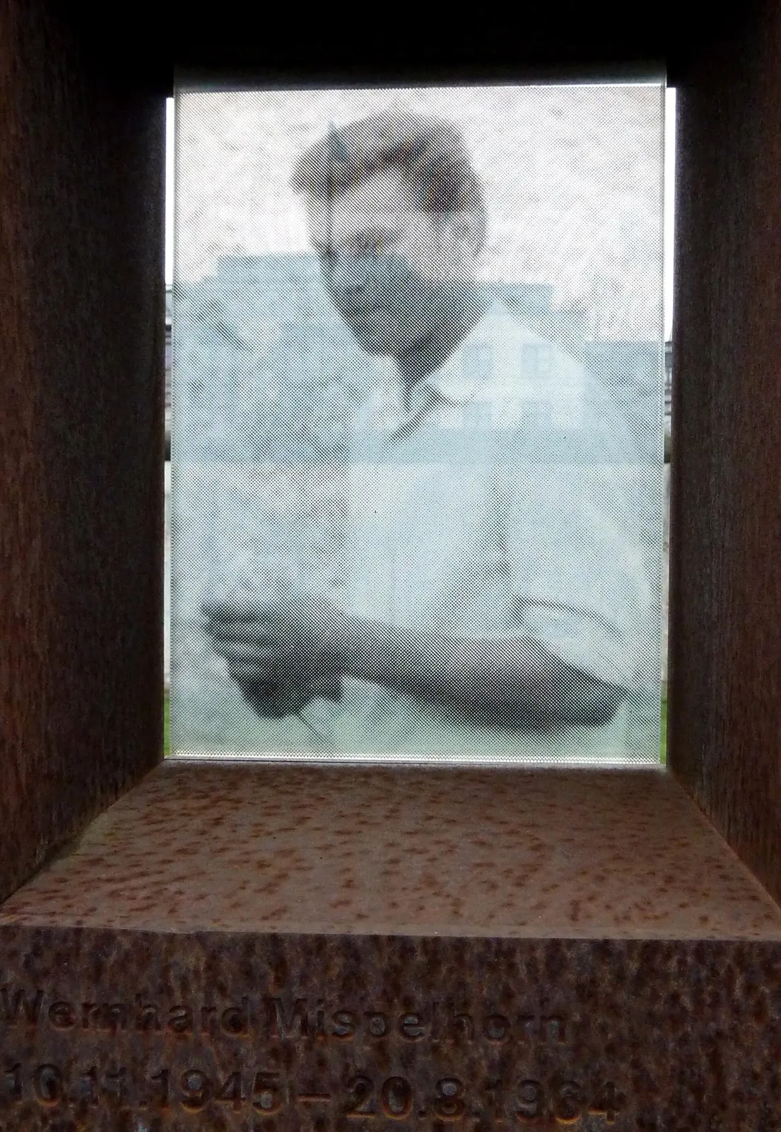 Faded black-and-white photograph of a young man in a light shirt, set in a rusted steel niche engraved 'Wernhard Mispelhorn 10.11.1945 – 20.8.1964'