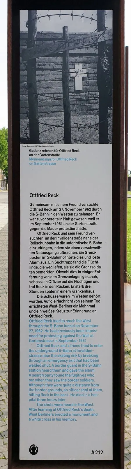 Tall rusted-steel panel with a black-and-white photograph of a wooden memorial sign and a cross behind barbed wire, above a German and English text headed 'Ottfried Reck'