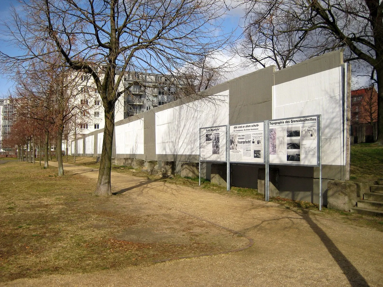 A long section of white-painted concrete wall with three information boards in front of it stands on a dry lawn among bare trees; apartment and office buildings rise behind.