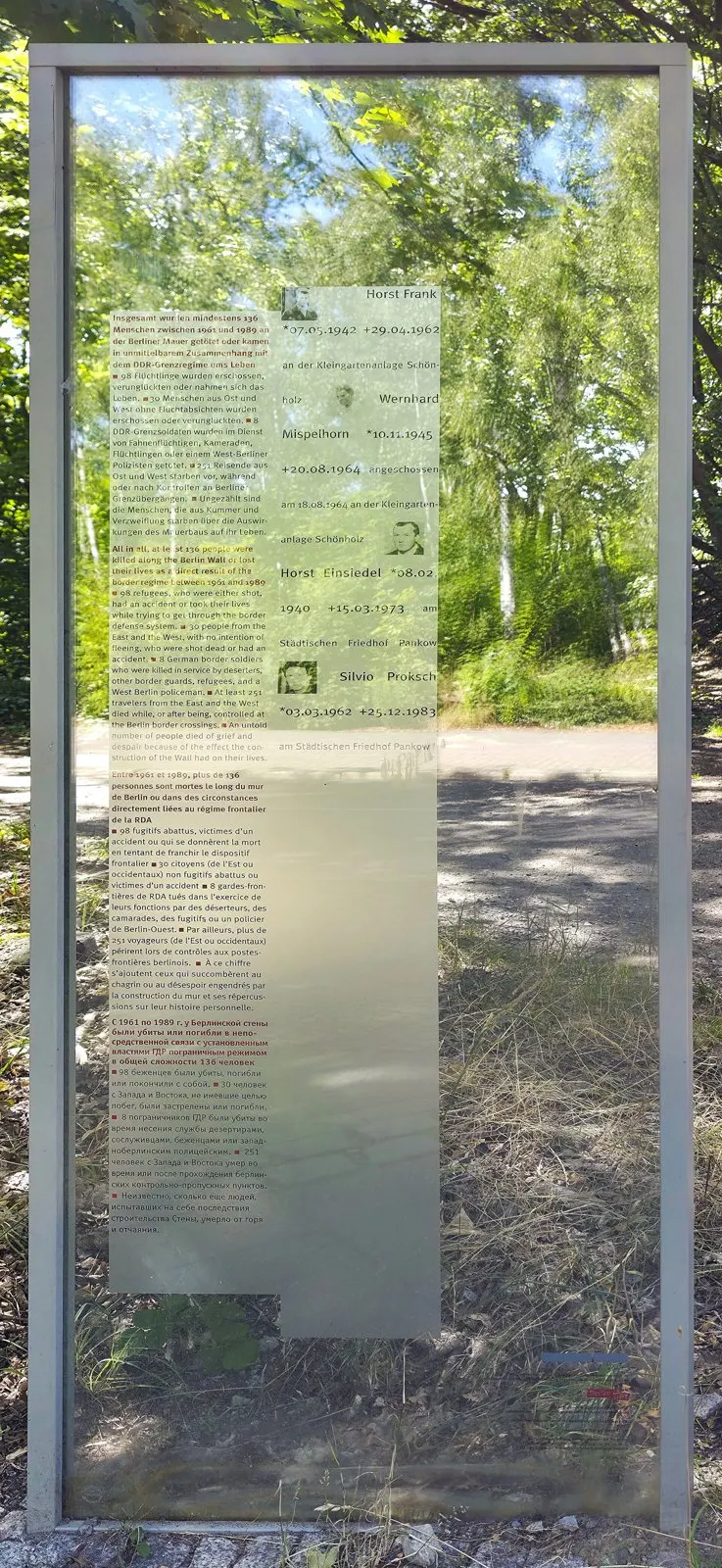 Tall glass panel with printed text and small portrait photographs in a steel frame, trees visible through the glass