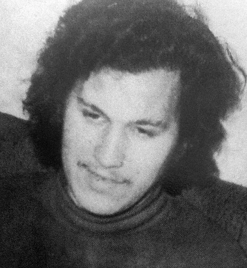 Black-and-white photograph of a young man with long dark curly hair and a roll-neck sweater, looking down