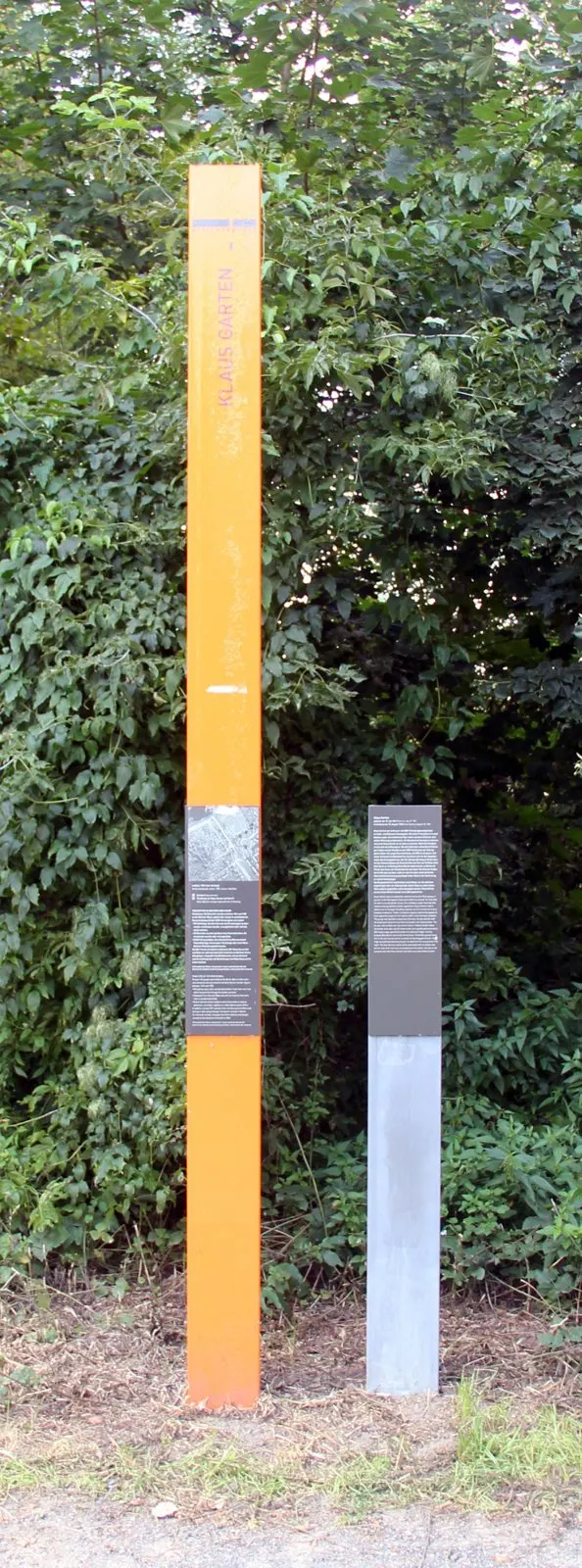 Tall orange column bearing the name "Klaus Garten" beside a grey panel with text, in front of dense green bushes