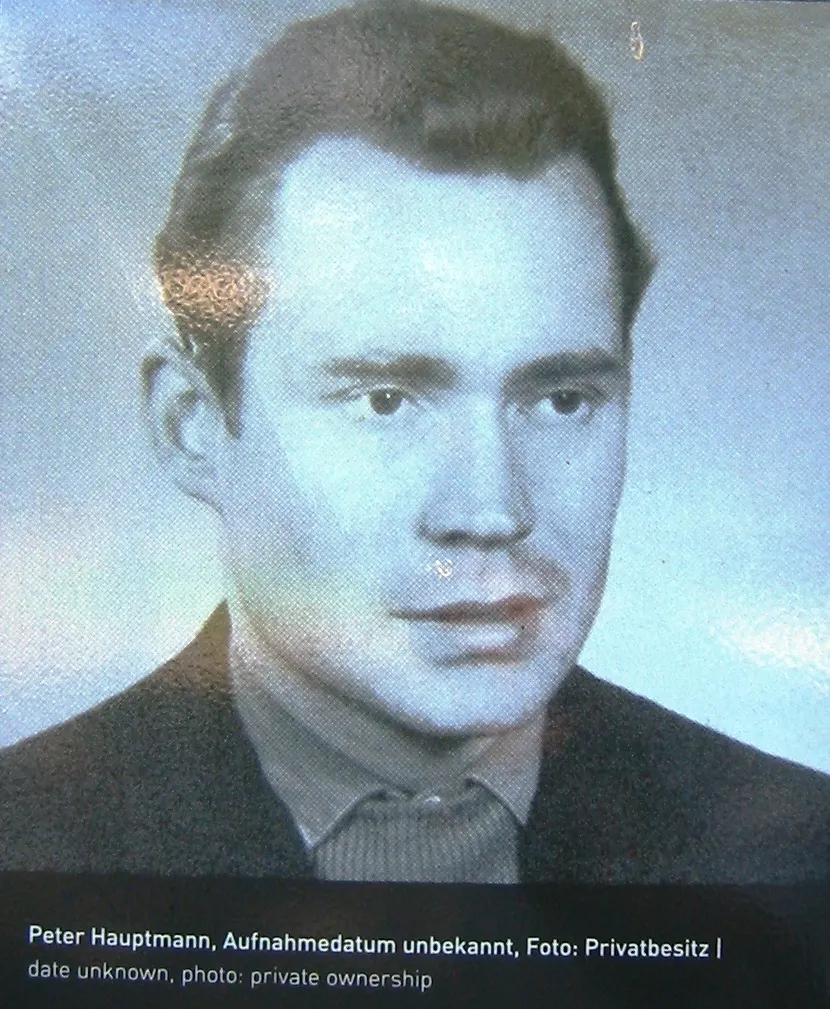Black-and-white portrait photograph of a man with swept-back dark hair, a dark jacket and a patterned shirt, with a printed caption below