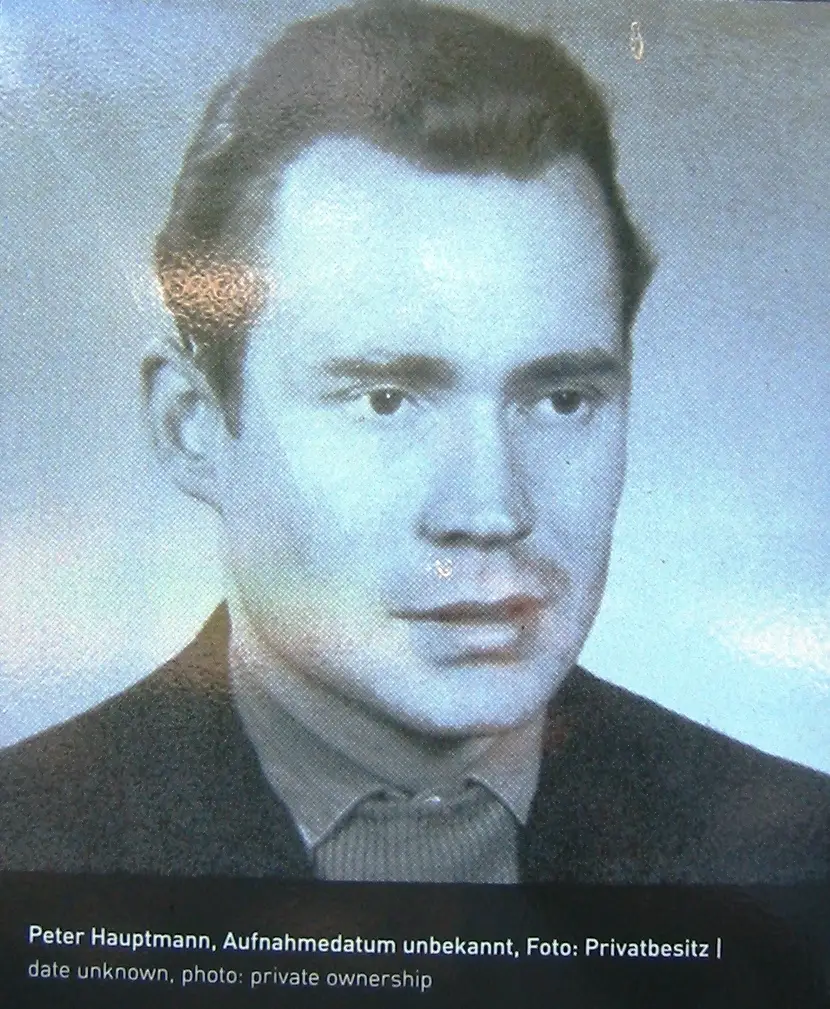 Black-and-white portrait photograph of a man with swept-back dark hair, a dark jacket and a patterned shirt, with a printed caption below