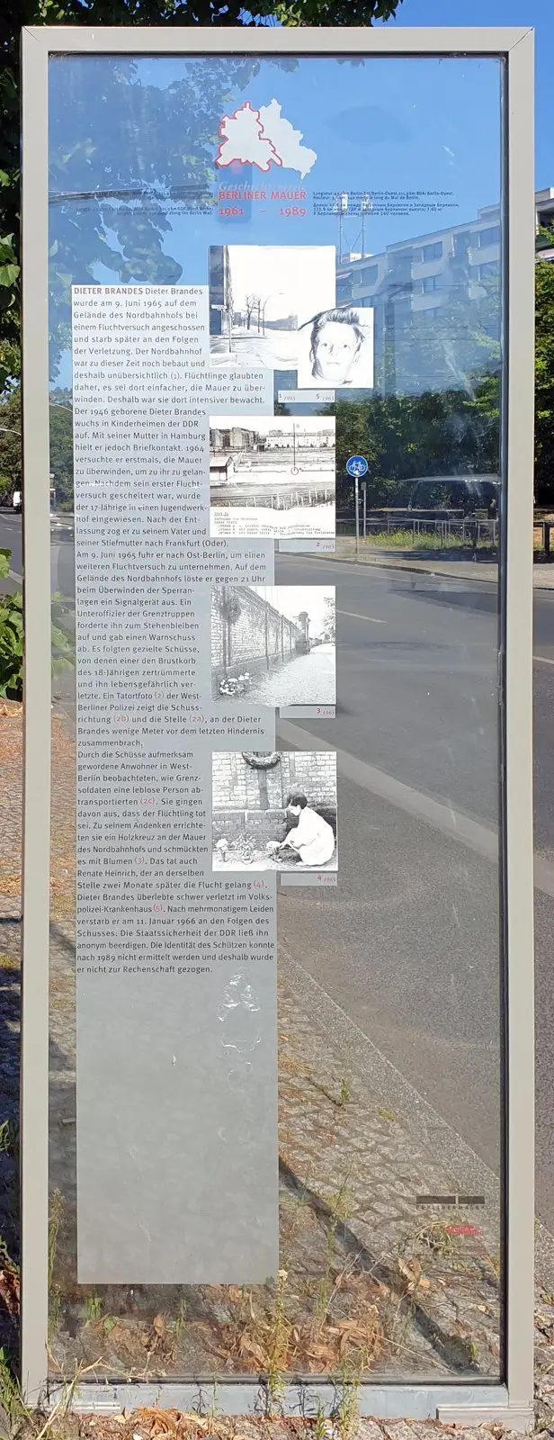 Tall glass panel in a steel frame with German text and small black-and-white photographs, including a portrait of a young man, beside a road