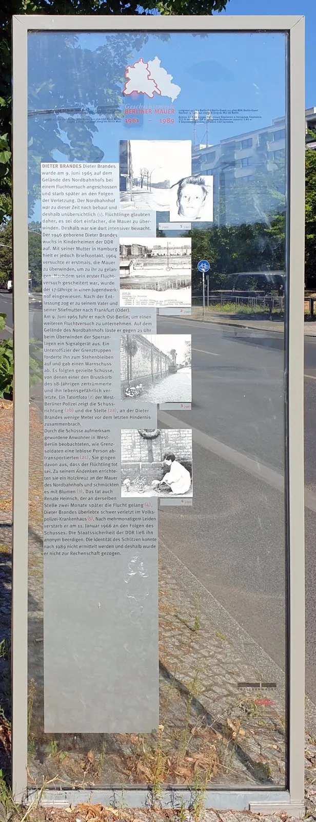 Tall glass panel in a steel frame with German text and small black-and-white photographs, including a portrait of a young man, beside a road