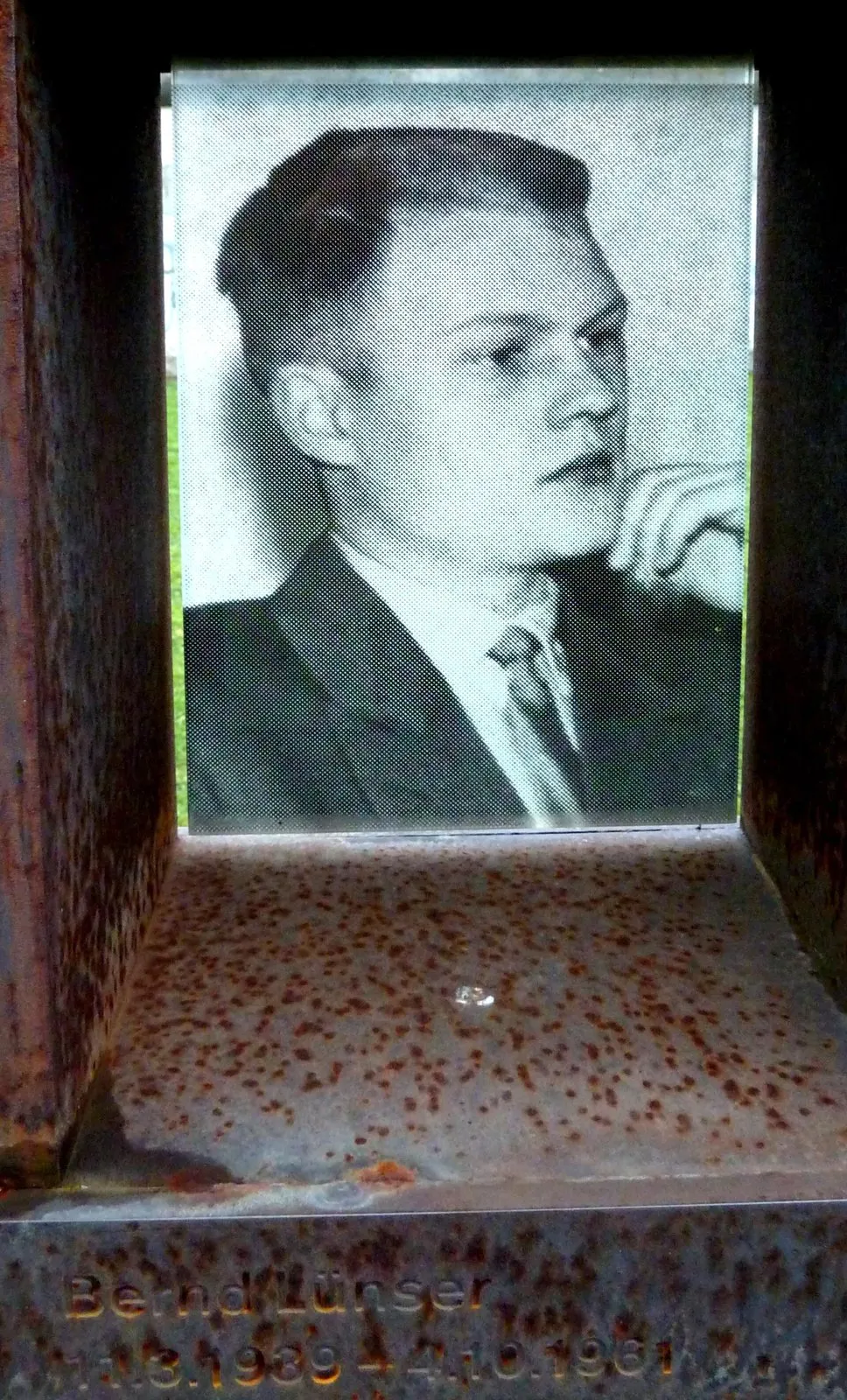 Black-and-white photograph of a young man in a suit and tie, hand at his chin, behind glass in a rusted steel niche