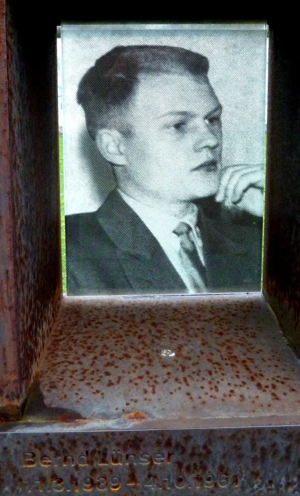 Black-and-white photograph of a young man in a suit and tie, hand at his chin, behind glass in a rusted steel niche