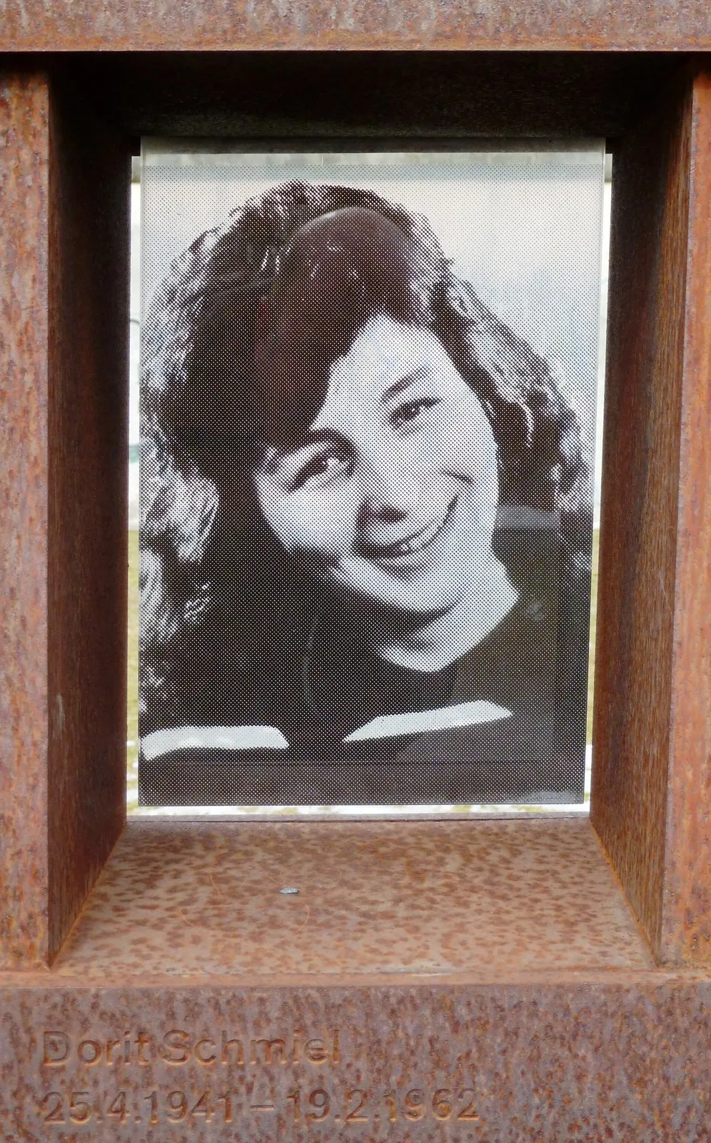 Black-and-white photograph of a smiling young woman with long curly hair behind glass in a rusted steel niche, her name engraved below