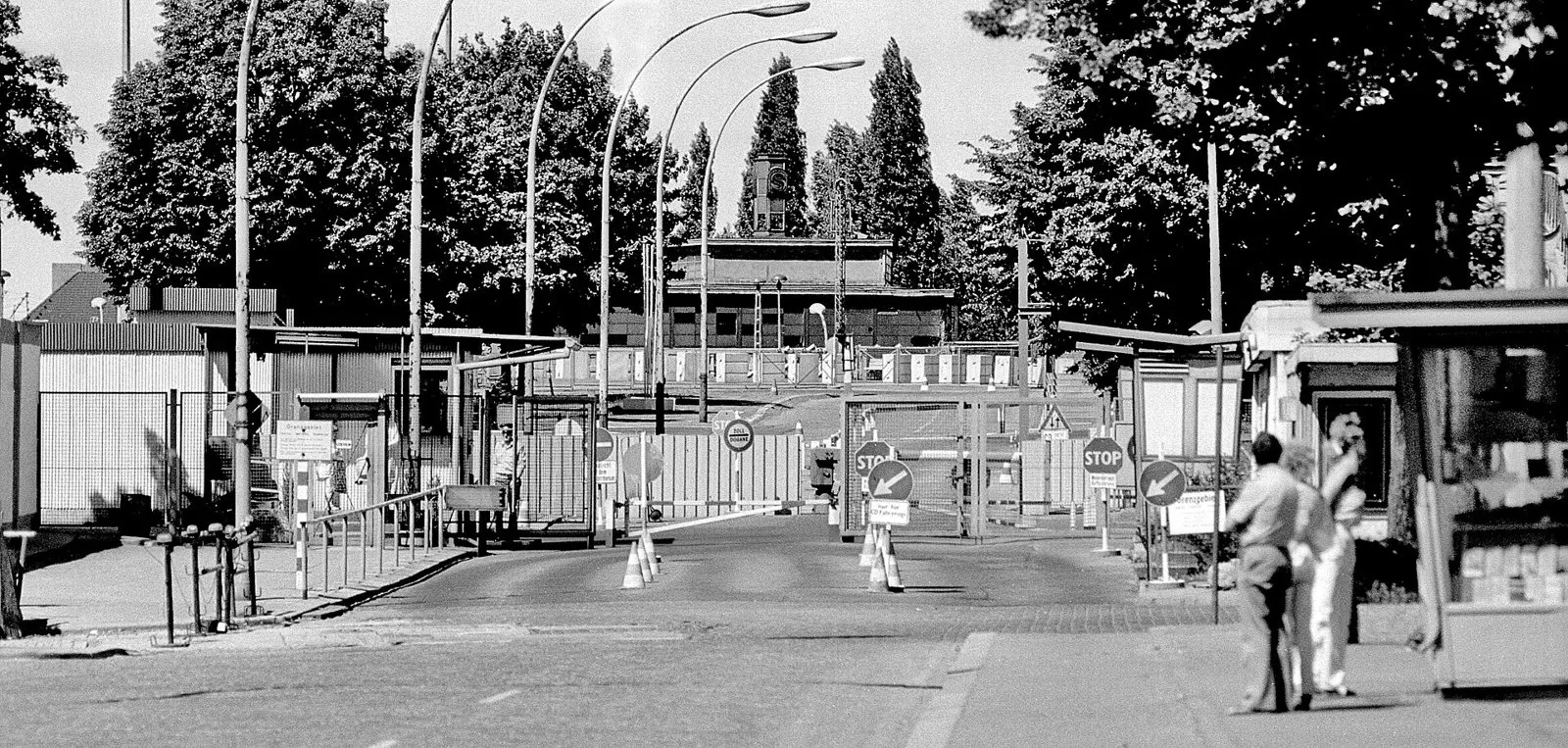 Black-and-white photograph of a border checkpoint: a road with barriers, stop signs and a row of curved street lamps leads towards low buildings and tall poplars; two people stand beside a kiosk on the right.