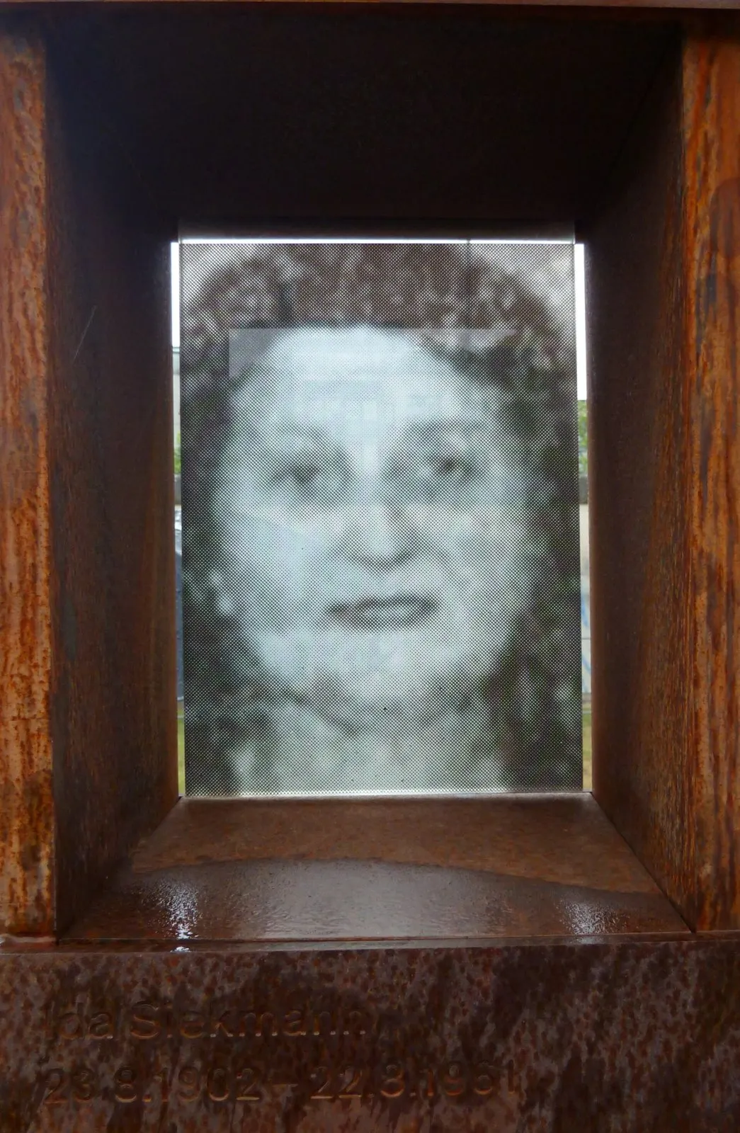 Black-and-white photograph of a woman with dark curly hair, set behind glass in a rusted steel niche with her name engraved below